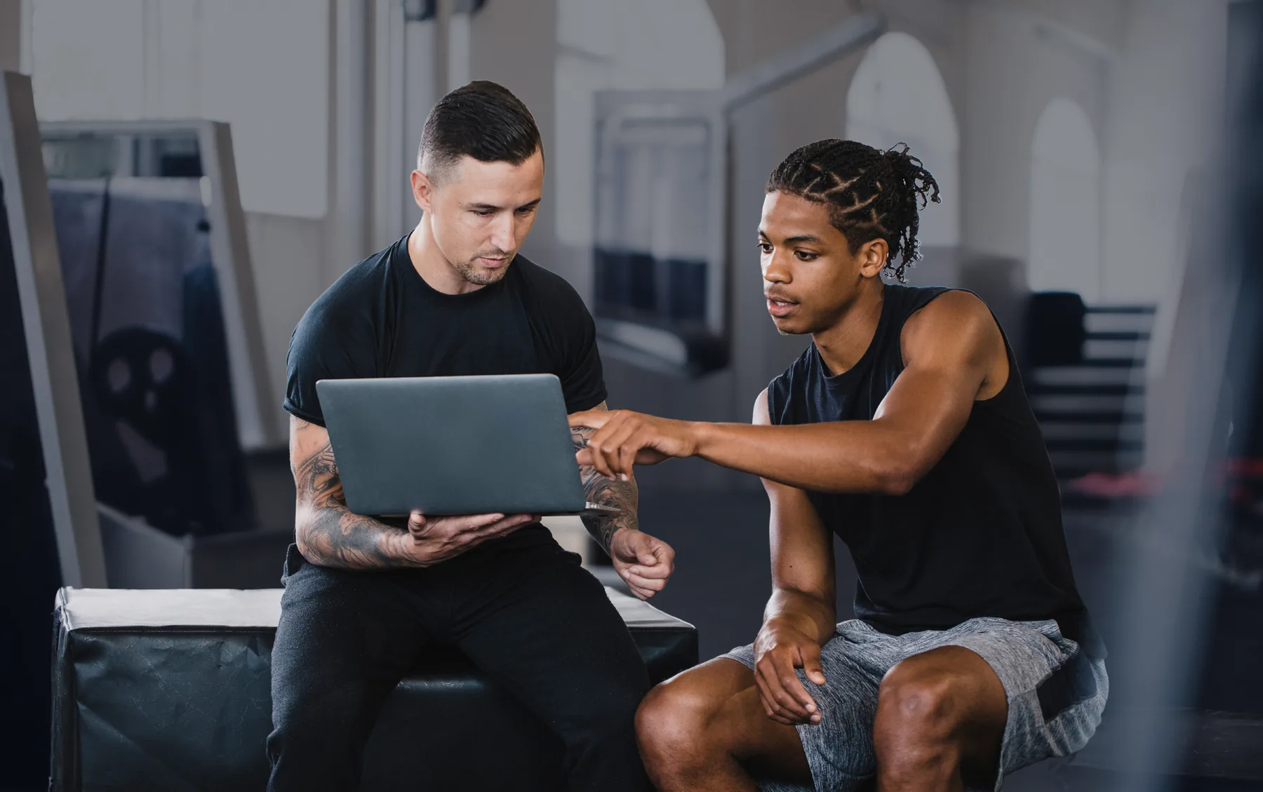 A trainer and a member sitting on a plyo box in the gym, looking at the member's readings on a laptop